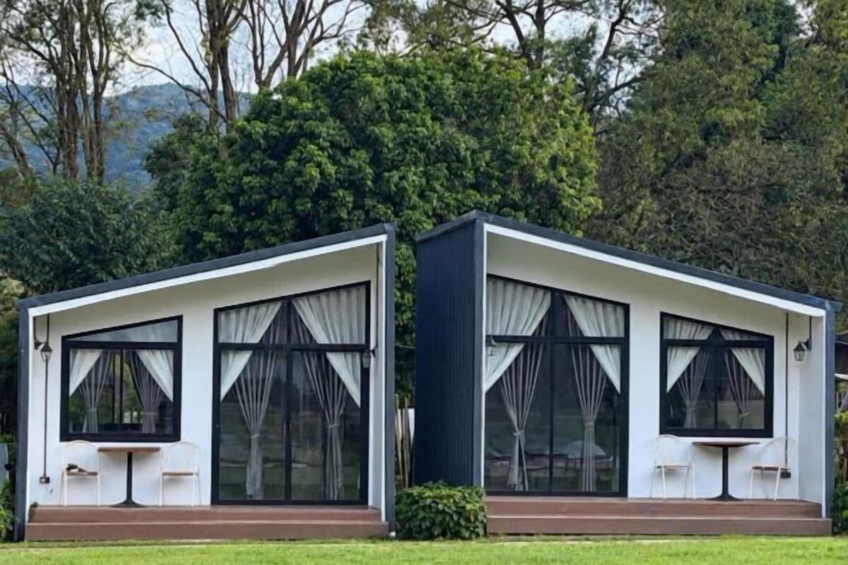 Deux bâtiments modernes, blancs, de plain-pied, dotés de grandes fenêtres et d’un toit plat, nichés dans un environnement luxuriant et verdoyant.