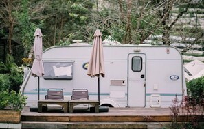 A white RV with two chairs and two umbrellas, parked outdoors with greenery in the background.