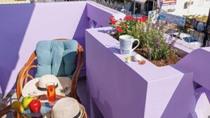 A balcony with a purple wall, a wicker chair, a hat, a glass of drink, and a small plant with red flowers. - Thena apartment IN the city center (Limenas Chersonisou)