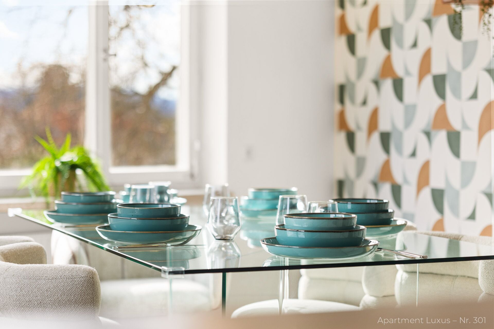 Ein Esstisch mit Glasplatte und blauen Tellern, platziert in einer modernen Wohnung mit geometrischer Tapete und Blick auf Bäume.