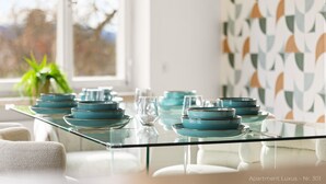 Ein Esstisch mit Glasplatte und blauen Tellern, platziert in einer modernen Wohnung mit geometrischer Tapete und Blick auf Bäume.