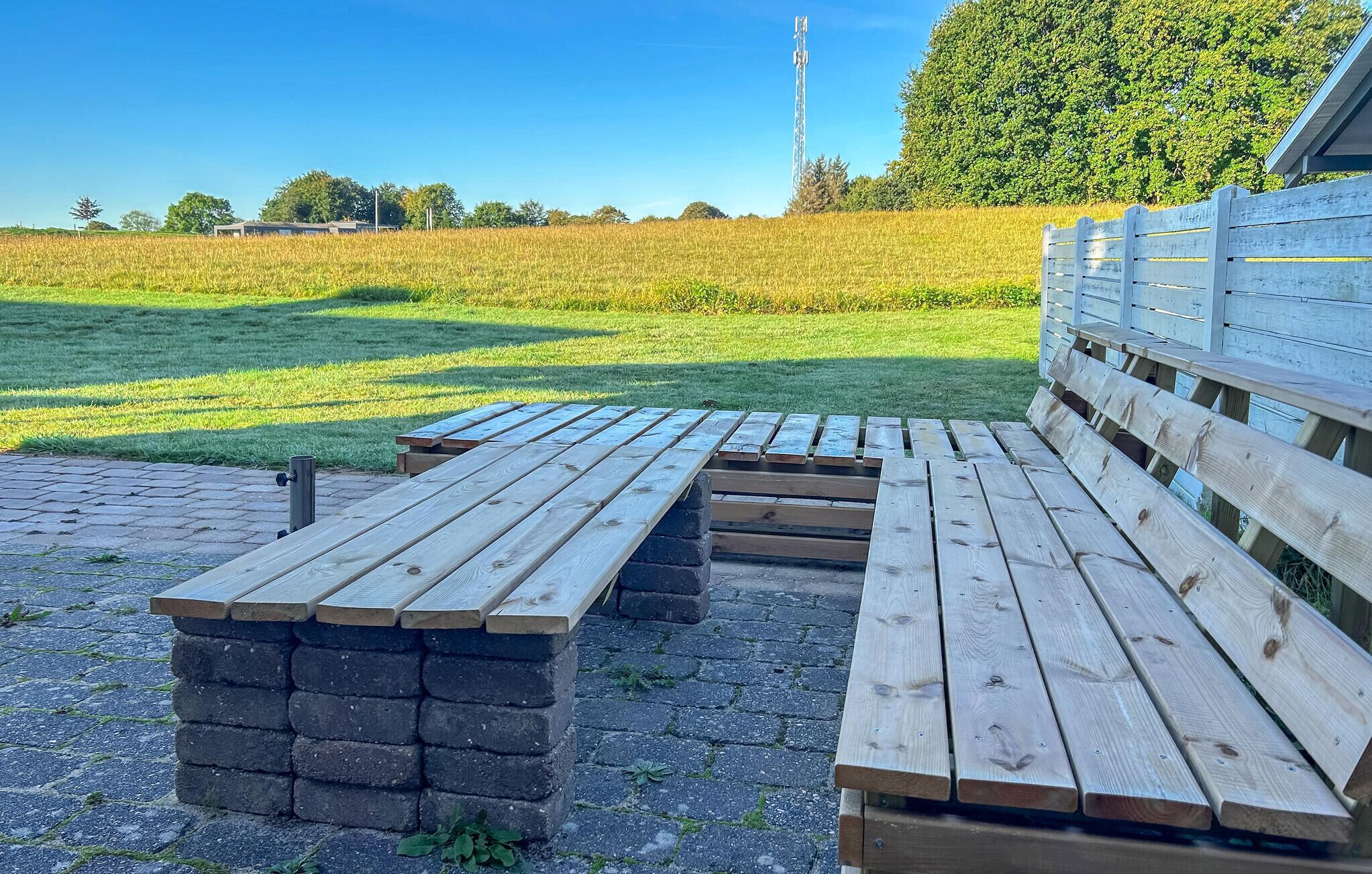 Une table de pique-nique avec des bancs et un socle en briques, installée sur une surface pavée, avec un champ d’herbe et des arbres en arrière-plan.