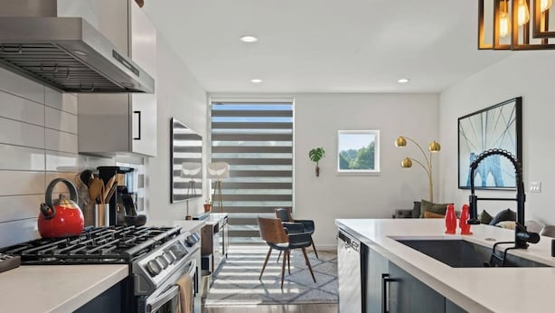 Uma cozinha moderna com fogão, forno e máquina de lavar loiça. Existe uma área de refeições com cadeiras e mesa. Uma janela proporciona luz natural.
