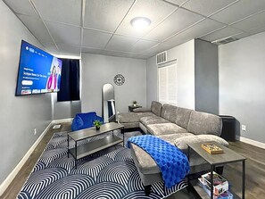Ein modernes Wohnzimmer mit einem Ecksofa, einem flachbildfernseher an der Wand, einer Uhr und einem gemusterten Teppich.