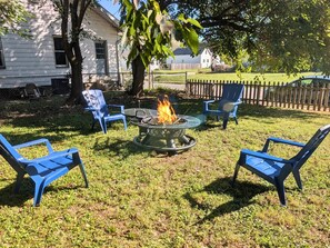 Ein Garten mit einem Feuerkorb, umgeben von blauen Stühlen, einem Haus im Hintergrund und einem Holzzaun.