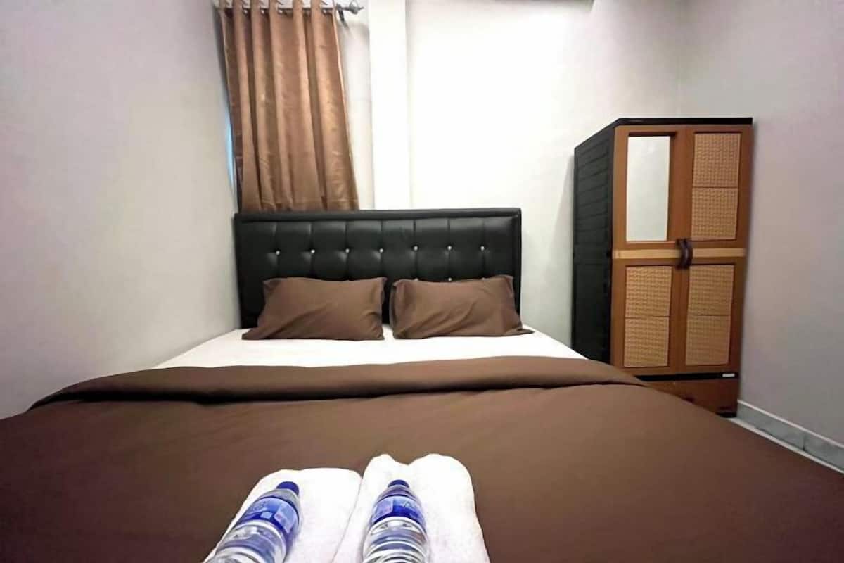 Ein ordentlich bezogenes Bett mit brauner Tagesdecke und Kissen, ein hölzerner Kleiderschrank mit Spiegel und ein Fenster mit braunen Vorhängen.