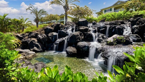 Una cascata che si getta su rocce in una piscina limpida, circondata da una vegetazione rigogliosa e un edificio sullo sfondo.