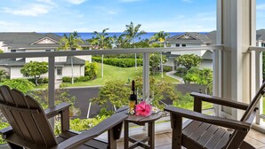 Un balcone con due poltrone Adirondack, un tavolino con una bottiglia e un fiore, e vista su una zona residenziale con case e un campo da golf.