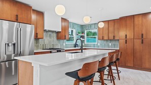 Eine moderne Küche mit Holzausstattung, einem Edelstahlkühlschrank, einer großen Kochinsel inklusive Barhocker und Pendelleuchten.