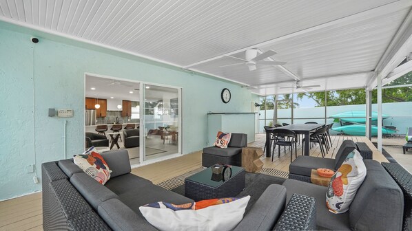 A covered outdoor seating area with sofas and a dining table, a kitchen visible through a glass door, and a clock on the wall.