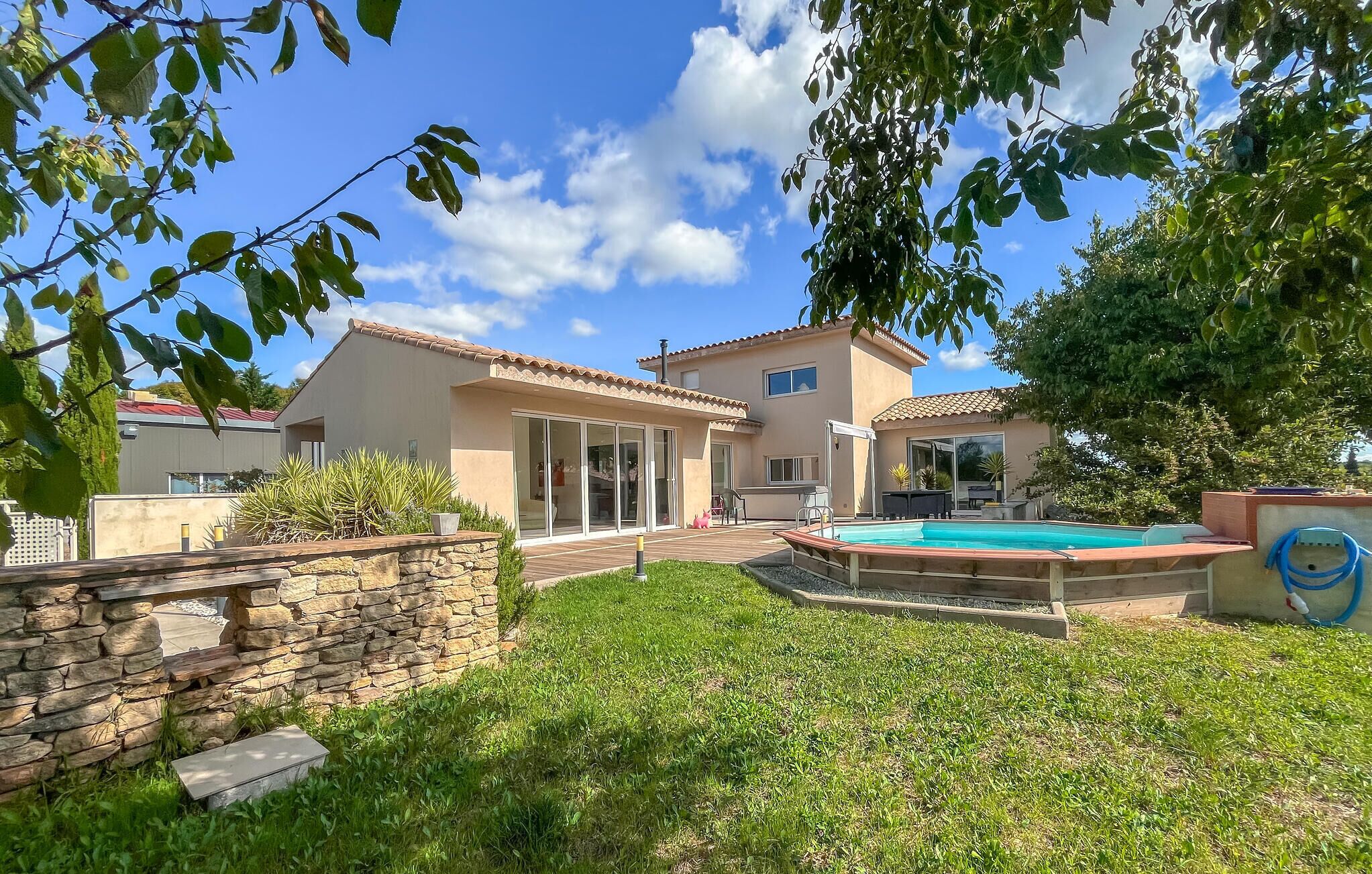 A two-story house with a swimming pool, stone wall, and a patio area.