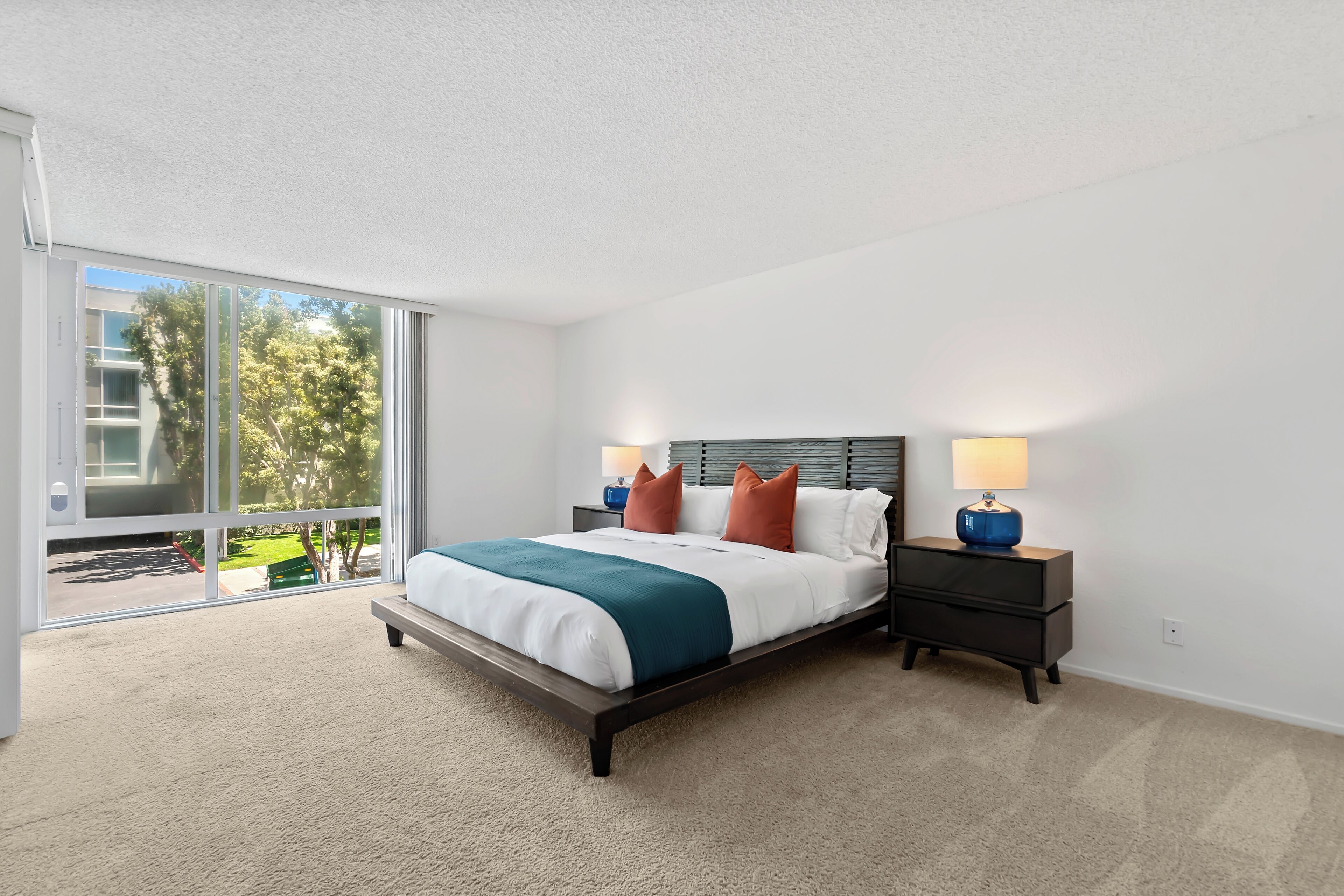 Quarto moderno com uma cama grande, duas mesas de cabeceira com abajures, e vista para um gramado verde e um edifício através de uma porta de vidro deslizante.
