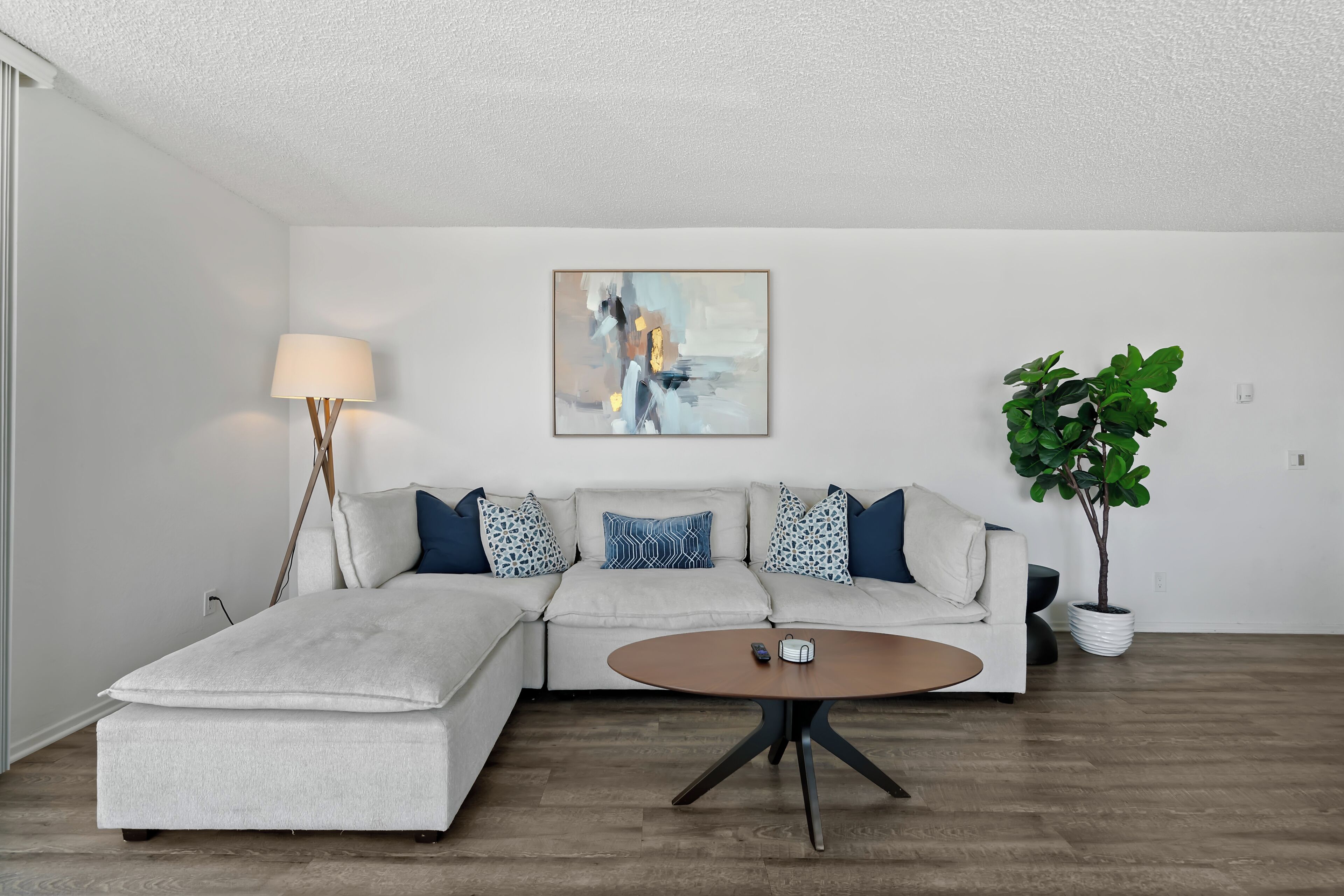 A modern living room with a sectional sofa, a round coffee table, a floor lamp, a painting on the wall, and a potted plant.