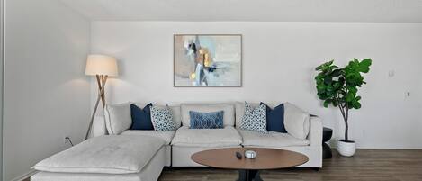 A modern living room with a sectional sofa, a round coffee table, a floor lamp, a painting on the wall, and a potted plant.