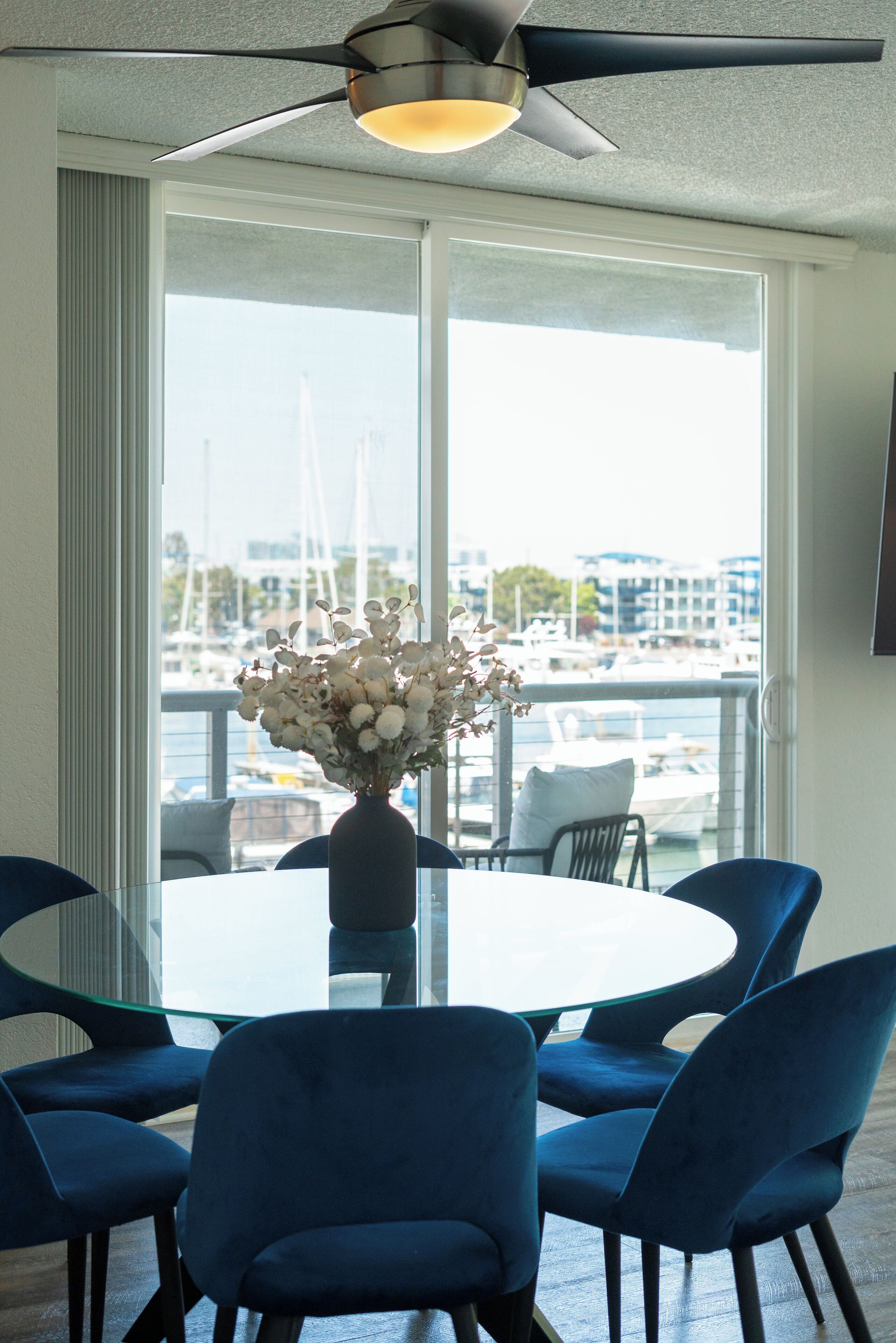 A dining area with a glass table, blue chairs, a vase with flowers, and a view of a marina with boats.
