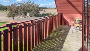 En træterrasse med rødt gelænder, et bord med stole, samt udsigt over et mark og træer.