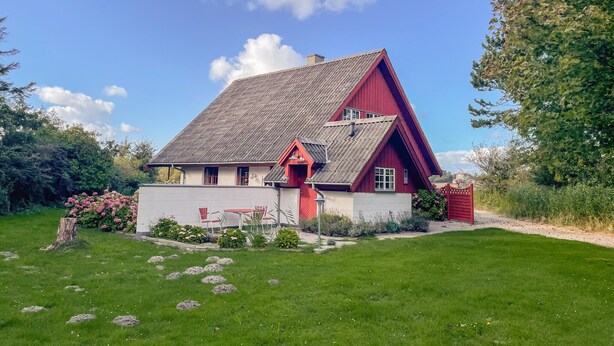 Et rødt og hvidt hus med skrå tag, omgivet af en velplejet græsplæne og blomstrende planter.