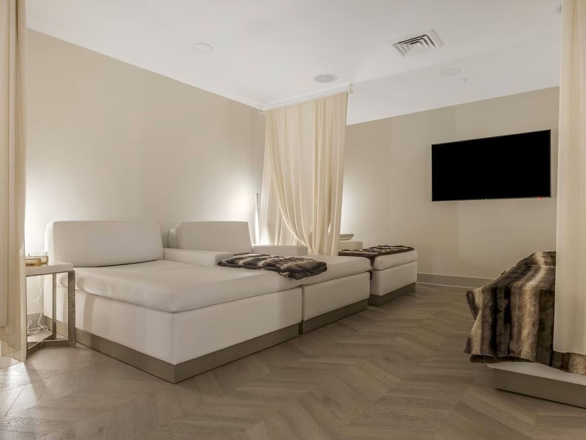 Una habitación de hotel moderna con una cama grande, un sofá más pequeño, un televisor montado en la pared y piso de madera.