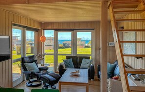 Un salon chaleureux avec un décor intérieur en bois, de grandes fenêtres offrant une vue sur la mer et les bâtiments environnants, un coin salon confortable avec une table, et une échelle en bois.