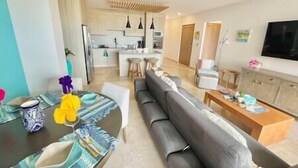A modern living room with a black sofa, a dining table with a glass top, and a kitchen in the background.