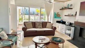 A modern living room with a brown sofa, two glass-top tables, a kitchen area with a white countertop, and a fireplace.