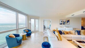 A spacious living area with a large window overlooking the sea, a white sofa with gold and blue cushions, and a blue armchair.
