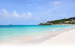 Ein Strand mit klarem türkisblauem Wasser, weißem Sand und Gebäuden, die an einem Hügel im Hintergrund liegen.