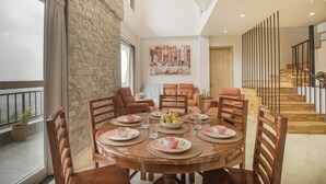 Un coin repas avec une table ronde dressée pour six personnes, entourée de chaises en bois. On aperçoit un mur en pierre et un escalier avec une rampe au fond.