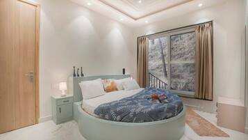 Un dormitorio con una cama curva, una mesita de noche, una lámpara de noche, una ventana con cortinas y vistas a un bosque.