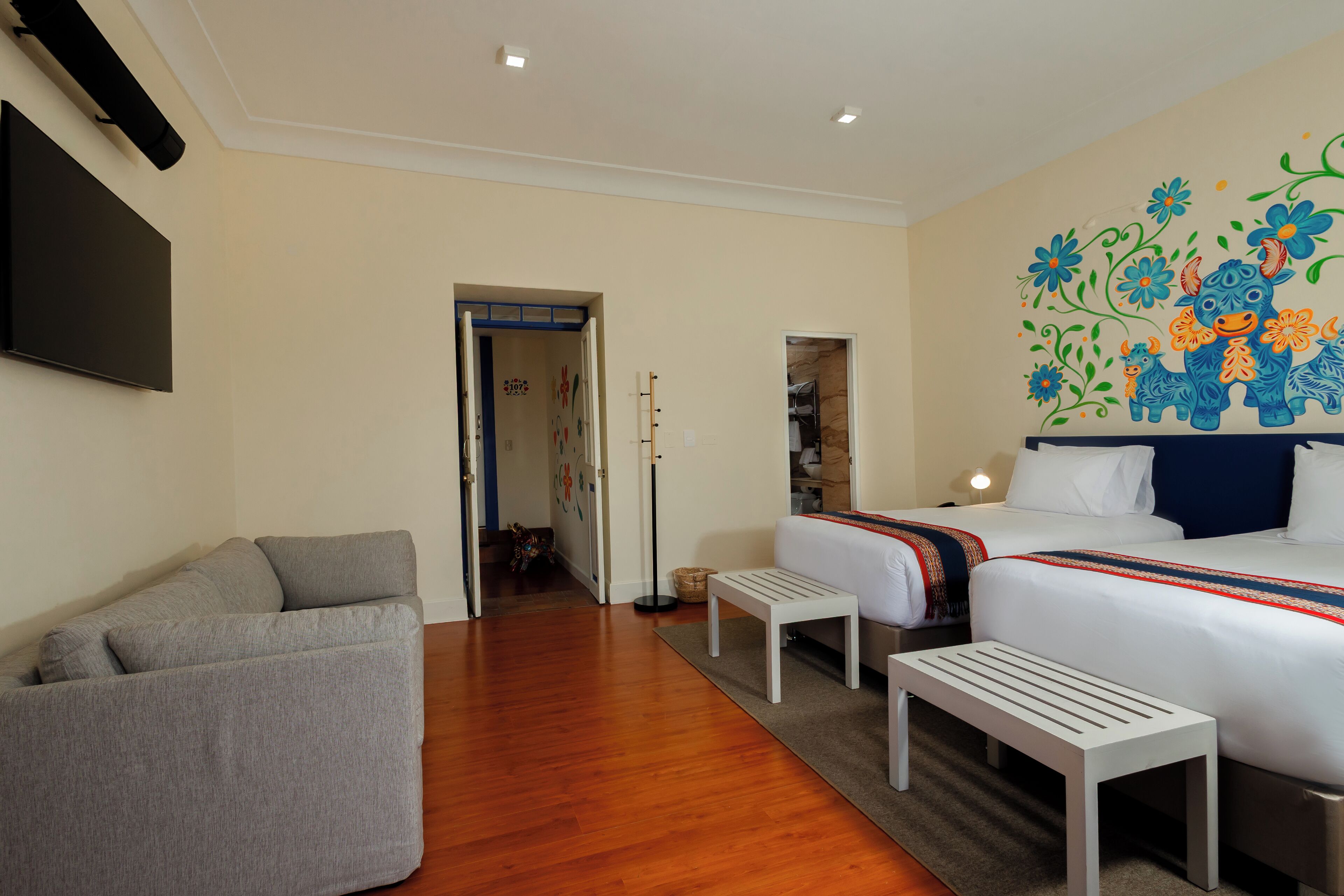 A hotel room with a sofa, two beds, a TV, and a colorful wall mural.