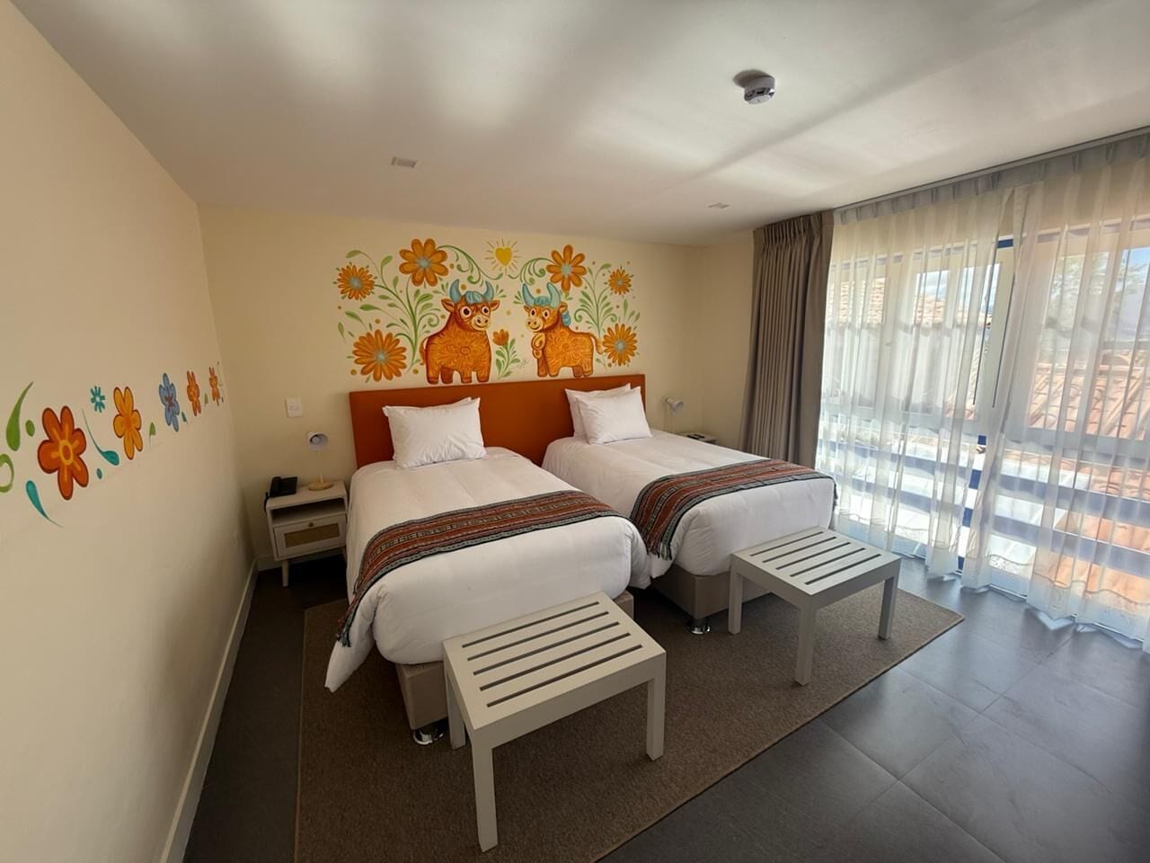 A hotel room with two beds, a nightstand, a small table, and a window with sheer curtains.