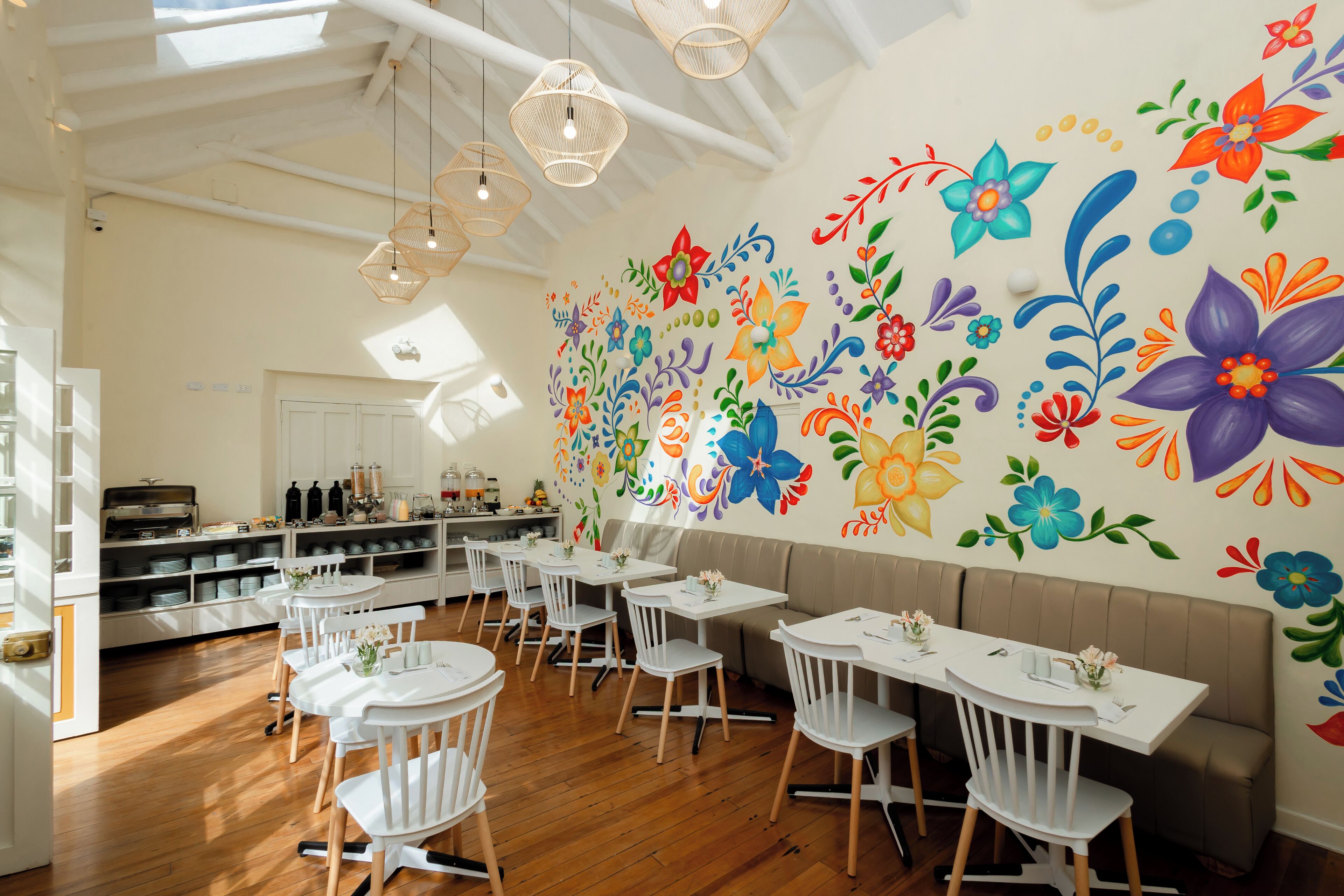 A restaurant with white tables and chairs, a wooden floor, and a colorful floral mural on the wall.