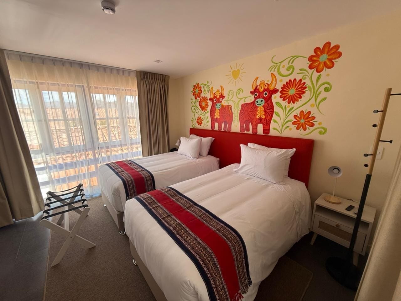 A hotel room with two beds, a red headboard, a window with curtains, a desk with a chair, and a wall mural.