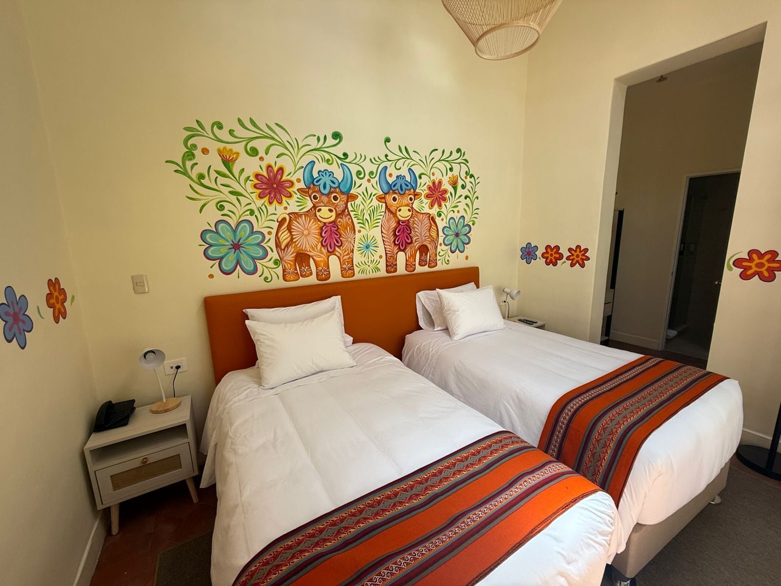 Ein Hotelzimmer mit zwei Betten, einem Nachttisch, einer Lampe und einer Wandmalerei mit bunten Blumen und Tieren.