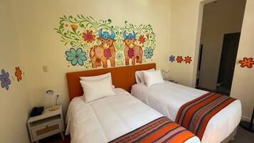 Habitación de hotel con dos camas, una mesita de noche, una lámpara y un mural en la pared con flores y animales de colores.