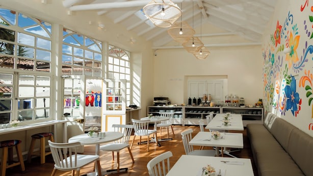 A bright, open dining area with white tables and chairs, large windows, and a colorful mural on the wall. - 7 Toritos Boutique Hotel (Cusco)