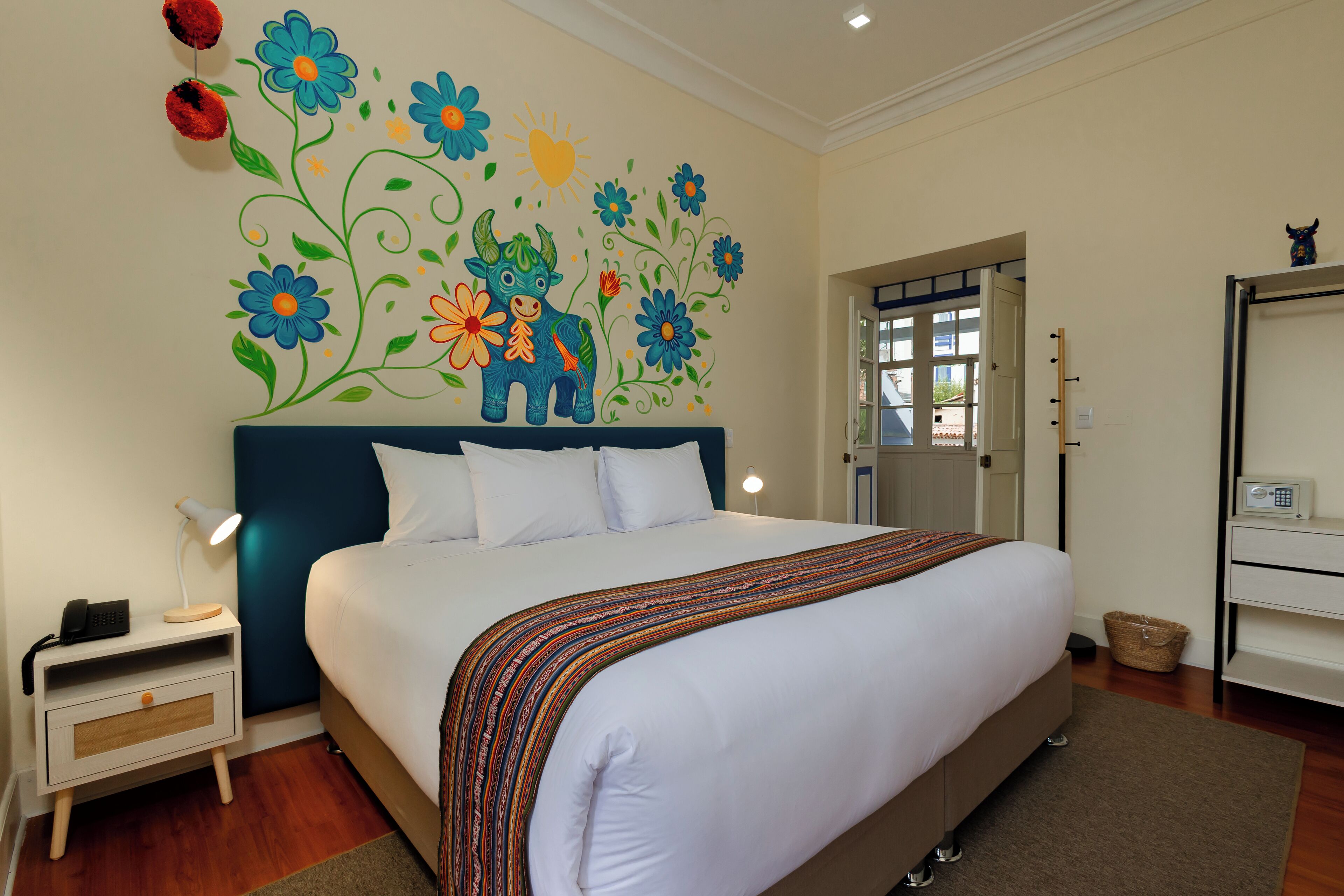 Quarto com uma cama grande, uma mesa de cabeceira com abajur, um mural na parede e uma porta que dá para outro cômodo.