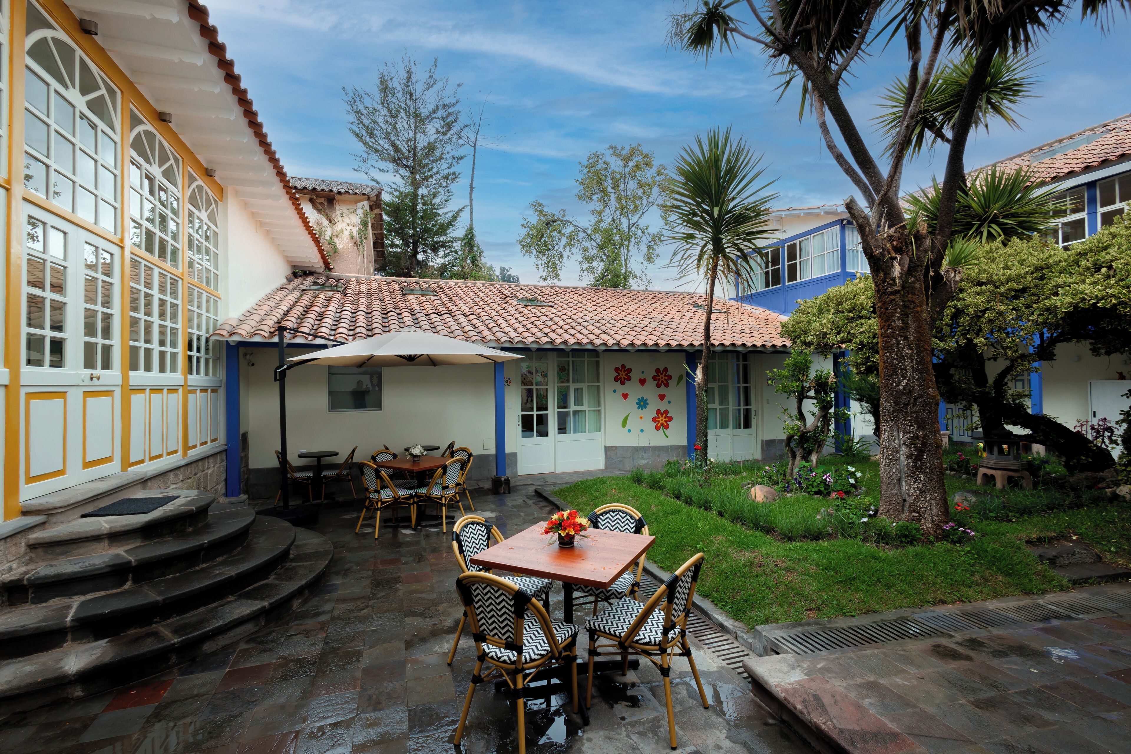 Eine Terrasse mit Tisch und Stühlen, ein überdachter Sitzbereich und eine Treppe, die zu einem Gebäude mit großen Fenstern und einem Ziegeldach führt.