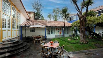 Eine Terrasse mit Tisch und Stühlen, ein überdachter Sitzbereich und eine Treppe, die zu einem Gebäude mit großen Fenstern und einem Ziegeldach führt.