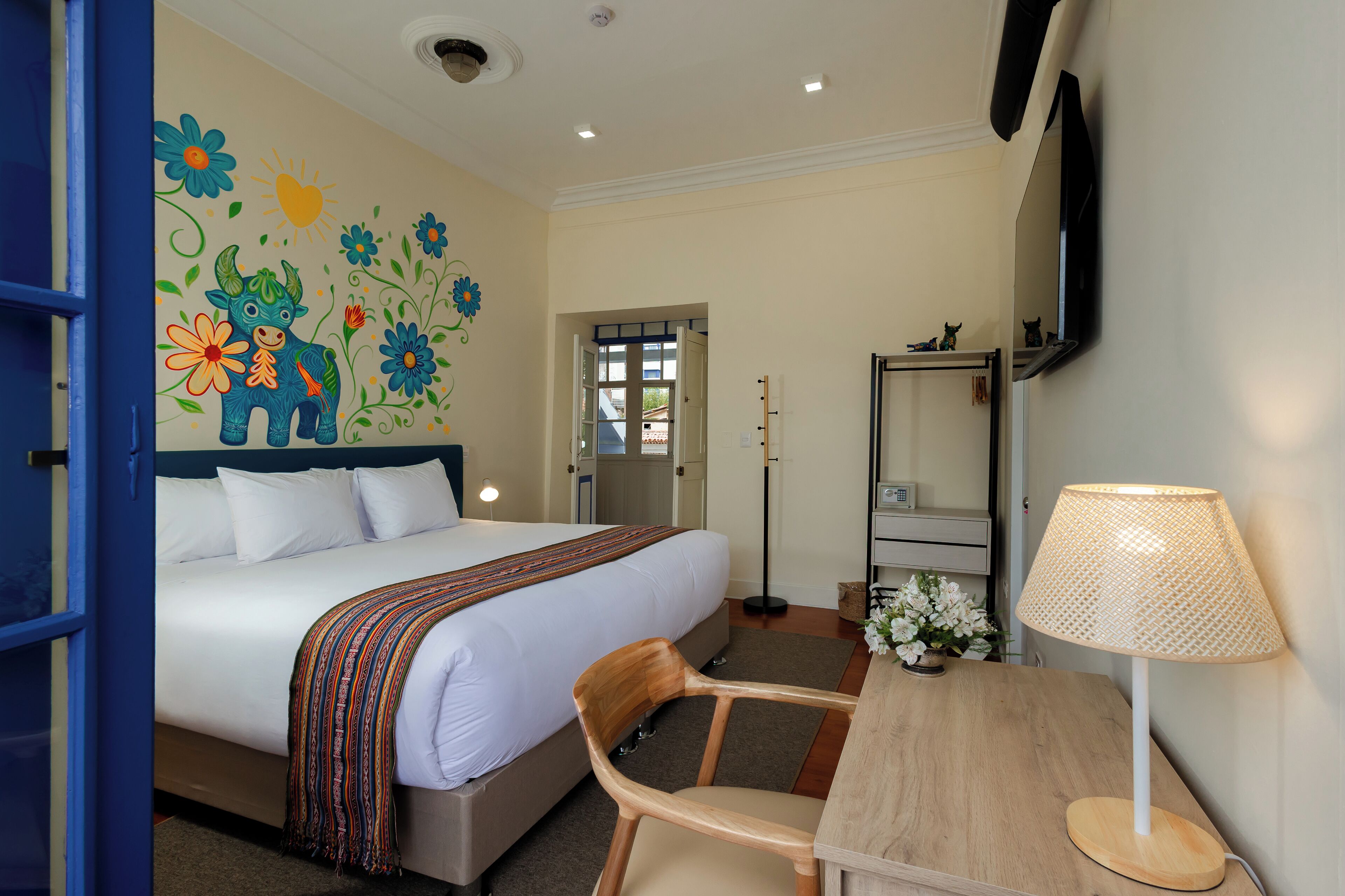 A hotel room with a bed, a desk with a lamp, a chair, a TV mounted on the wall, and a colorful mural on the wall.