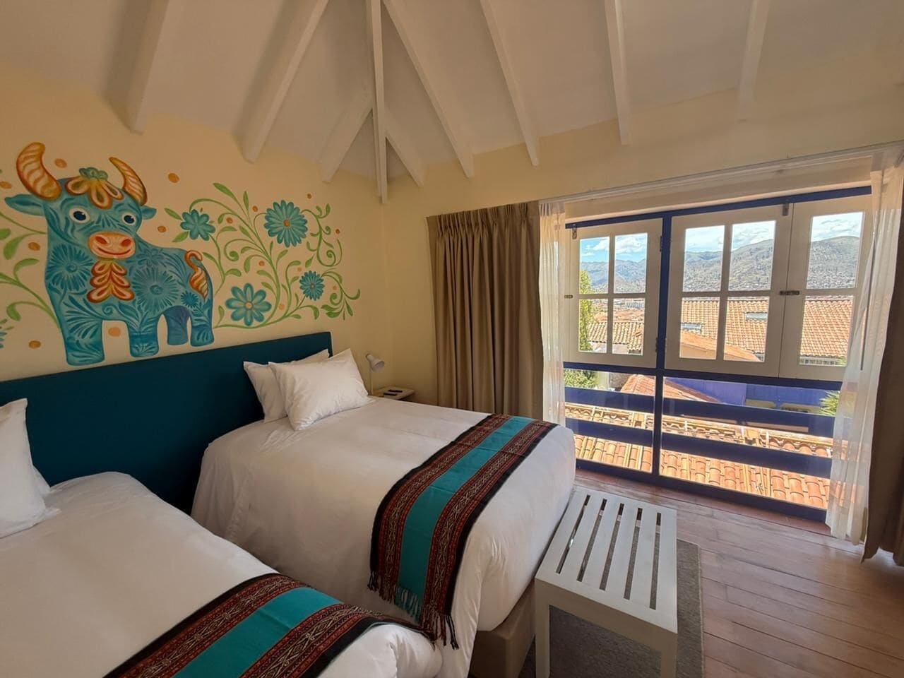 A hotel room with two beds, a balcony with a view of the city, and a mural on the wall.