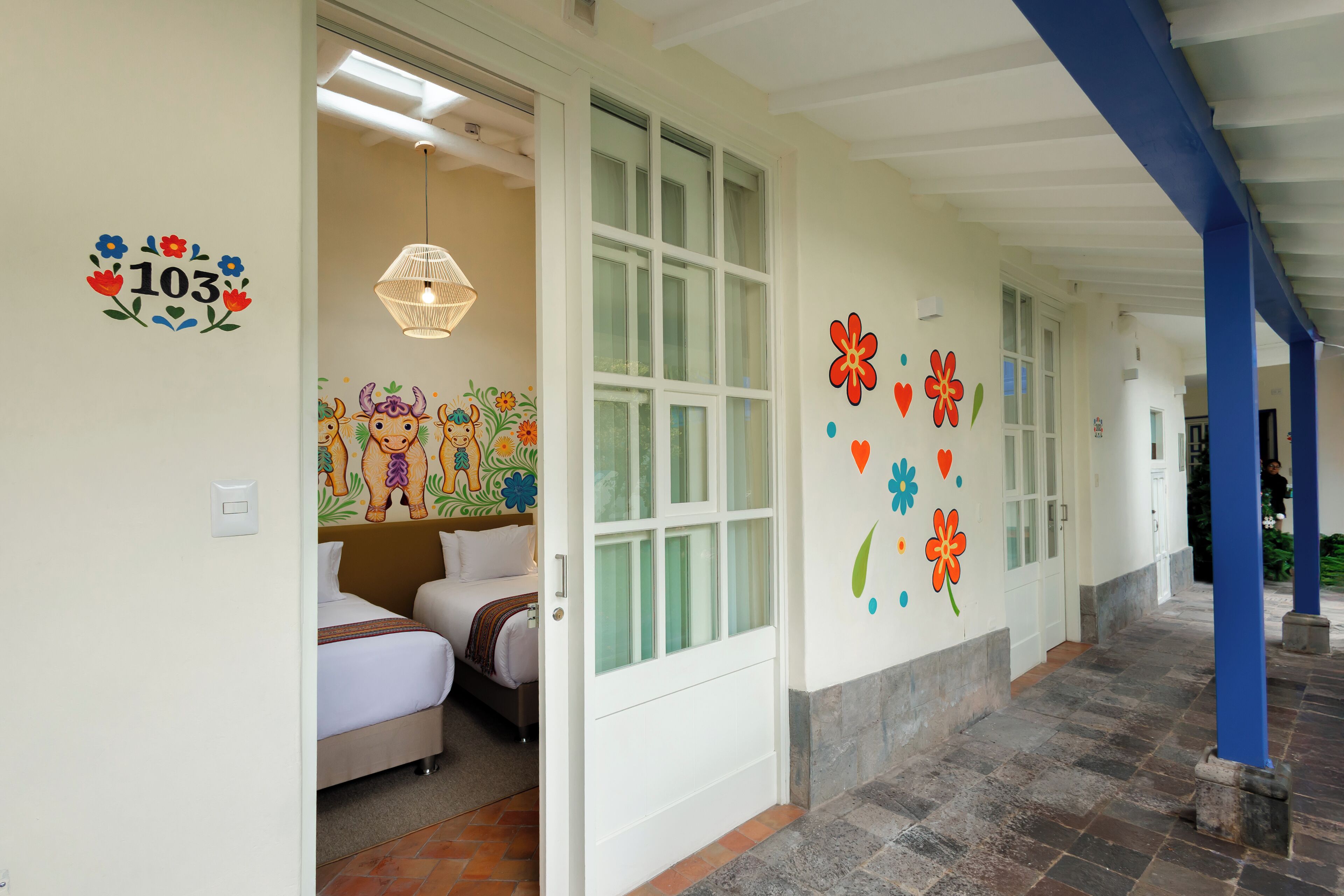 A hotel corridor with a bed, a door, and a wall with number 103 and floral decorations.