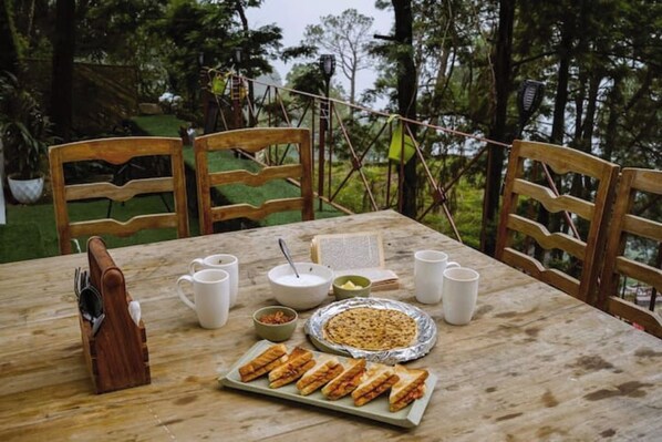 Una mesa de madera al aire libre con comida, incluyendo sándwiches, un tazón de sopa y una fuente de pan.