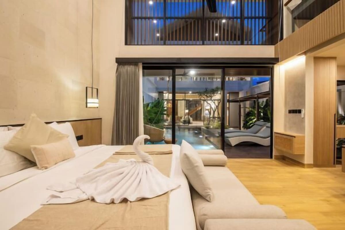 A modern hotel room with a bed, a sofa, a glass door leading to a pool area, and a view of the outdoors.