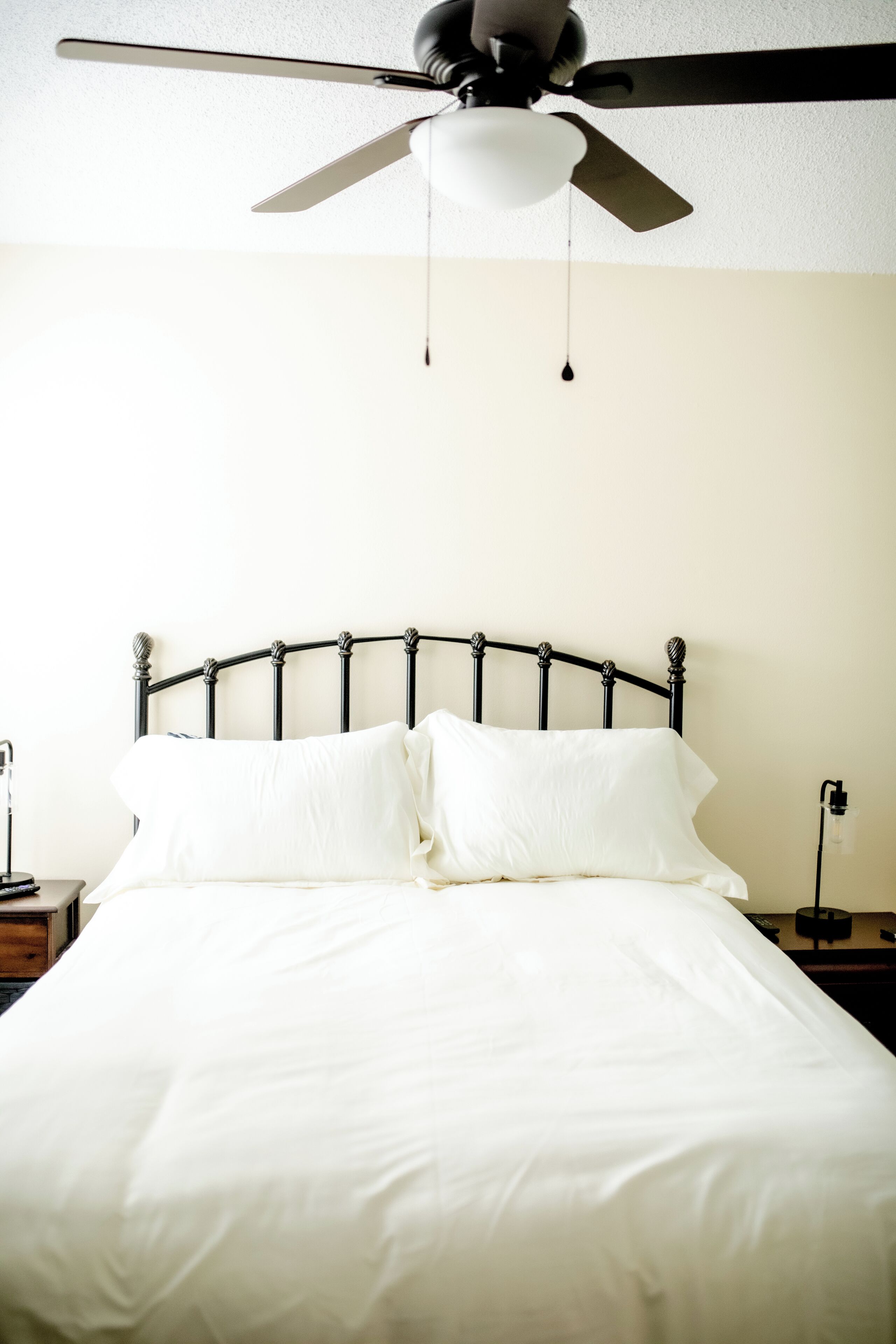 Een bed met een witte dekbedovertrek en kussens, een plafondventilator en een nachtkastje met een lamp.