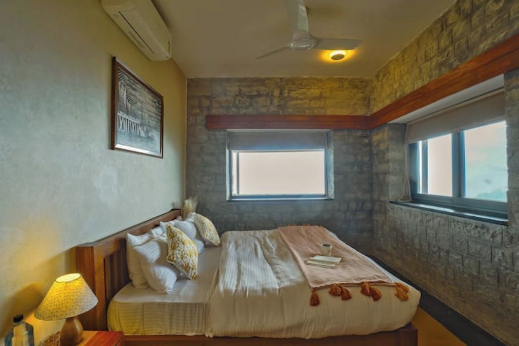 A bedroom with a stone wall, a bed with pillows, a window, and a ceiling fan.