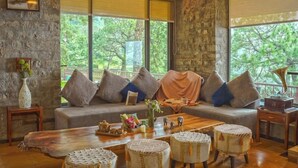 Una sala de estar acogedora con una pared de piedra, un ventanal con vistas a árboles, un sofá con cojines, una mesa de centro de madera y otomanas de mimbre.