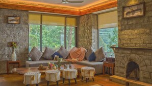 Una sala de estar acogedora con una chimenea de piedra, un sofá con cojines, una mesa de centro con adornos y vistas a zonas verdes a través de amplios ventanales.