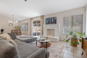 A modern living room with a grey sofa, a round coffee table, a fireplace, and a large window with blinds. - All-inclusive beautiful 3-bedroom home in NW Visalia (Visalia)