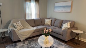 Ein Wohnzimmer mit einem grauen Ecksofa, einem runden Couchtisch mit Blumenvase und einem Gemälde an der Wand.
